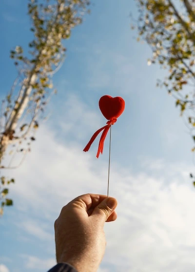 Kurs – Buddhismus – Liebe und Mitgefuehl Ein kleines, hübsches, rotes Herz mit einer roten Schleife, das gegen den Himmel gehalten wird.