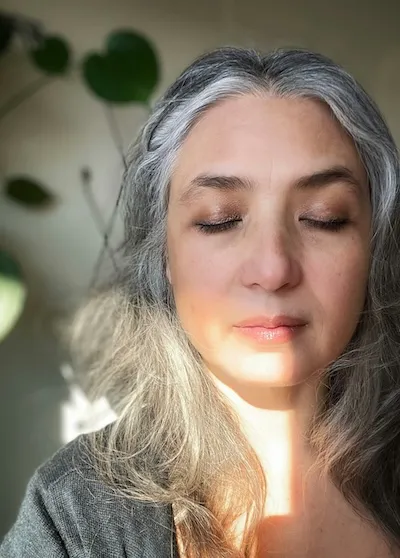 Meditation Basel – Der Mittlere Weg Ein Foto einer Frau mit geschlossenen Augen in meditierender Haltung, deren Gesicht teilweise von der Sonne beschienen wird, ohne dass es sie blendet.