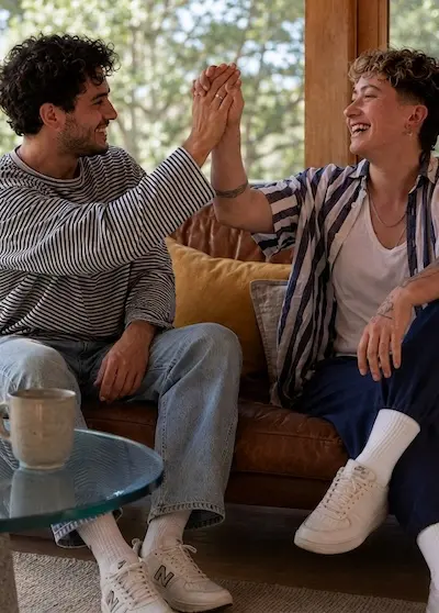 Schuldzuweisung loslassen Ein gut gelauntes Paar sitzt auf einem Sofa und gibt sich einen versöhnlichen High-Five Handschlag